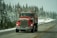 A red semi-truck with visible exhaust pipes is driving down a snowy road lined with evergreen trees. The truck features a message in white text on the front. The overcast sky and snow on the ground create a wintry atmosphere.