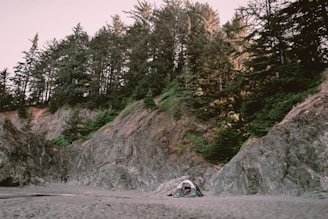 A serene campsite is nestled on a sandy beach surrounded by rocky cliffs and dense forest. A small green tent is pitched at the base of the cliffs, highlighting the contrast between the soft sand and the rugged natural terrain. Tall trees rise above the cliffs, adding depth and a natural canopy to the scene.