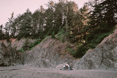 A serene campsite is nestled on a sandy beach surrounded by rocky cliffs and dense forest. A small green tent is pitched at the base of the cliffs, highlighting the contrast between the soft sand and the rugged natural terrain. Tall trees rise above the cliffs, adding depth and a natural canopy to the scene.