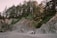 A serene campsite is nestled on a sandy beach surrounded by rocky cliffs and dense forest. A small green tent is pitched at the base of the cliffs, highlighting the contrast between the soft sand and the rugged natural terrain. Tall trees rise above the cliffs, adding depth and a natural canopy to the scene.