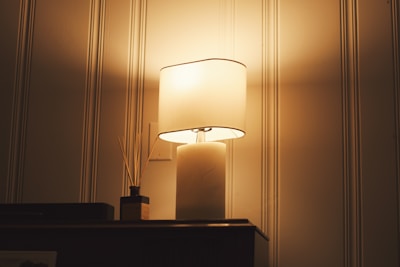 A warmly lit table lamp with a beige shade sits on top of a wooden piece of furniture. Next to the lamp, there is a small square decorative item with wooden sticks, possibly a reed diffuser. The background shows vertical striped wallpaper with a soft glow emanating from the lamp, casting a cozy ambiance.