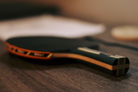 A table tennis paddle lies on a wooden surface, with a black rubber top and an orange trim. The background is blurred, giving focus to the paddle.