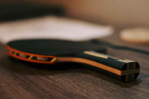 A table tennis paddle lies on a wooden surface, with a black rubber top and an orange trim. The background is blurred, giving focus to the paddle.