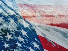 A close-up view of a textured fabric displaying the American flag pattern, featuring prominent stars and stripes. The colors appear slightly faded, suggesting a weathered or vintage look.