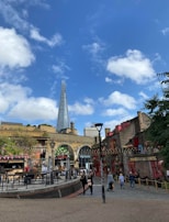 A courtyard with people walking and dining, surrounded by historic brick buildings. The tall, modern structure of The Shard prominently rises into the sky in the background. Strings of lights and colorful storefronts add to the vibrant atmosphere.