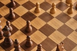 A chessboard with alternating light and dark brown squares is shown, featuring several wooden chess pieces. The pieces include pawns and other pieces forming a game setup but not arranged in a starting position. A combination of light-colored and dark-colored chess pieces are present.
