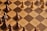 A chessboard with alternating light and dark brown squares is shown, featuring several wooden chess pieces. The pieces include pawns and other pieces forming a game setup but not arranged in a starting position. A combination of light-colored and dark-colored chess pieces are present.