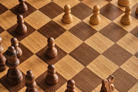 A chessboard with alternating light and dark brown squares is shown, featuring several wooden chess pieces. The pieces include pawns and other pieces forming a game setup but not arranged in a starting position. A combination of light-colored and dark-colored chess pieces are present.