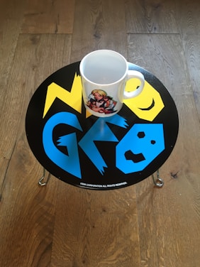 A white mug with a colorful illustration is placed on a small round table with a vibrant design. The tabletop features yellow and blue geometric shapes and letters on a black background. The table is set on a wooden floor with visible grain patterns.