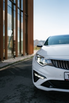 The image features the front section of a sleek, white car with its headlights on. The vehicle is parked on a paved road next to a modern building with tall glass windows reflecting the surrounding environment.