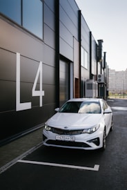 A white Kia car is parked next to a modern, dark-paneled building with large white letters displaying 'L4'. The weather appears clear, with reflections of clouds visible on the car's windshield.