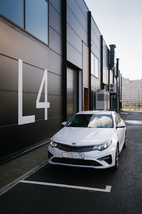 A white Kia car is parked next to a modern, dark-paneled building with large white letters displaying 'L4'. The weather appears clear, with reflections of clouds visible on the car's windshield.