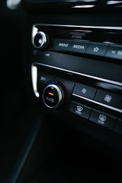 A close-up view of a car's dashboard featuring control buttons and dials for settings such as radio, media, and climate control. The central knob is labeled 'AUTO', with a red and blue color scheme indicating temperature control. The image includes labels like 'PASSENGER AIR BAG' and symbols for defrost settings.