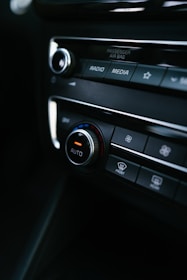A close-up view of a car's dashboard featuring control buttons and dials for settings such as radio, media, and climate control. The central knob is labeled 'AUTO', with a red and blue color scheme indicating temperature control. The image includes labels like 'PASSENGER AIR BAG' and symbols for defrost settings.