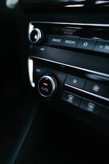 A close-up view of a car's dashboard featuring control buttons and dials for settings such as radio, media, and climate control. The central knob is labeled 'AUTO', with a red and blue color scheme indicating temperature control. The image includes labels like 'PASSENGER AIR BAG' and symbols for defrost settings.