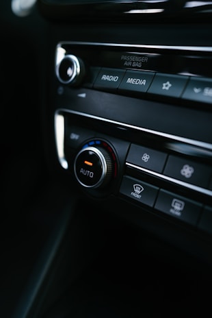A close-up view of a car's dashboard featuring control buttons and dials for settings such as radio, media, and climate control. The central knob is labeled 'AUTO', with a red and blue color scheme indicating temperature control. The image includes labels like 'PASSENGER AIR BAG' and symbols for defrost settings.