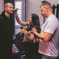 A woman applies makeup to a man's face while another man stands nearby holding a camera. The room appears to be a studio with a window in the background.