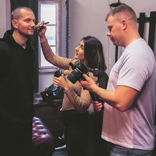 A woman applies makeup to a man's face while another man stands nearby holding a camera. The room appears to be a studio with a window in the background.