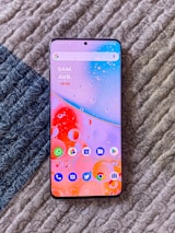 A smartphone with a vibrant and colorful wallpaper displaying various app icons is placed on a textured, patterned surface. The screen shows the time and date, and several unread notifications are visible on the apps.