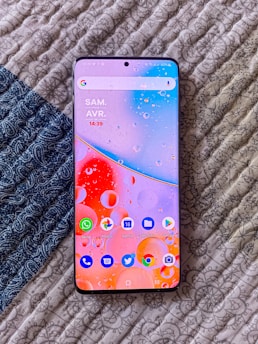 A smartphone with a vibrant and colorful wallpaper displaying various app icons is placed on a textured, patterned surface. The screen shows the time and date, and several unread notifications are visible on the apps.