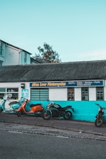 Several motorcycles are parked in front of a building with a sign reading 'Alan Lear Motorcycles'. The building is painted white and turquoise with large windows, and the sign also mentions services like MOTs, sales, service, and parts. The sky is clear and blue, suggesting a bright day.