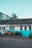 Several motorcycles are parked in front of a building with a sign reading 'Alan Lear Motorcycles'. The building is painted white and turquoise with large windows, and the sign also mentions services like MOTs, sales, service, and parts. The sky is clear and blue, suggesting a bright day.