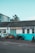 Several motorcycles are parked in front of a building with a sign reading 'Alan Lear Motorcycles'. The building is painted white and turquoise with large windows, and the sign also mentions services like MOTs, sales, service, and parts. The sky is clear and blue, suggesting a bright day.