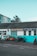Several motorcycles are parked in front of a building with a sign reading 'Alan Lear Motorcycles'. The building is painted white and turquoise with large windows, and the sign also mentions services like MOTs, sales, service, and parts. The sky is clear and blue, suggesting a bright day.
