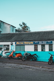 Several motorcycles are parked in front of a building with a sign reading 'Alan Lear Motorcycles'. The building is painted white and turquoise with large windows, and the sign also mentions services like MOTs, sales, service, and parts. The sky is clear and blue, suggesting a bright day.