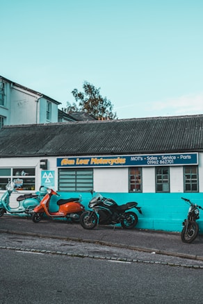 Several motorcycles are parked in front of a building with a sign reading 'Alan Lear Motorcycles'. The building is painted white and turquoise with large windows, and the sign also mentions services like MOTs, sales, service, and parts. The sky is clear and blue, suggesting a bright day.