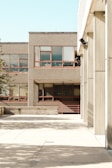 The photograph features an urban environment with multiple buildings displaying a minimalist architectural style characterized by concrete facades and rectangular windows. The open courtyard area is largely empty, with shadows from nearby structures casting patterns on the pavement. The scene is brightly lit, indicative of clear weather.