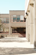 The photograph features an urban environment with multiple buildings displaying a minimalist architectural style characterized by concrete facades and rectangular windows. The open courtyard area is largely empty, with shadows from nearby structures casting patterns on the pavement. The scene is brightly lit, indicative of clear weather.