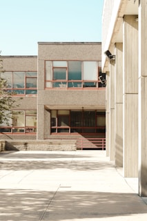 The photograph features an urban environment with multiple buildings displaying a minimalist architectural style characterized by concrete facades and rectangular windows. The open courtyard area is largely empty, with shadows from nearby structures casting patterns on the pavement. The scene is brightly lit, indicative of clear weather.