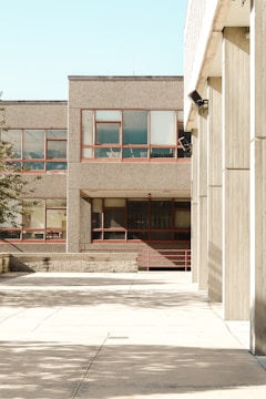 The photograph features an urban environment with multiple buildings displaying a minimalist architectural style characterized by concrete facades and rectangular windows. The open courtyard area is largely empty, with shadows from nearby structures casting patterns on the pavement. The scene is brightly lit, indicative of clear weather.