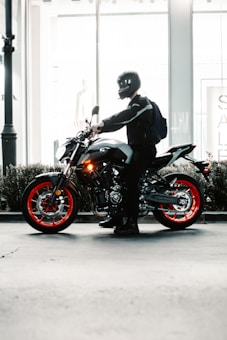 A motorcyclist, wearing a helmet and dark clothing, is posed next to a modern motorcycle with red-accented wheels. The setting is urban, possibly in front of a building with large glass windows, and there is signage visible in the background.