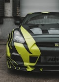 A close-up view of a sports car with a striking black and neon yellow design. The car features sleek, aerodynamic bodywork and is partially parked in front of a gray industrial garage door. The headlights are sharply defined, and the front grille showcases a distinctive logo.