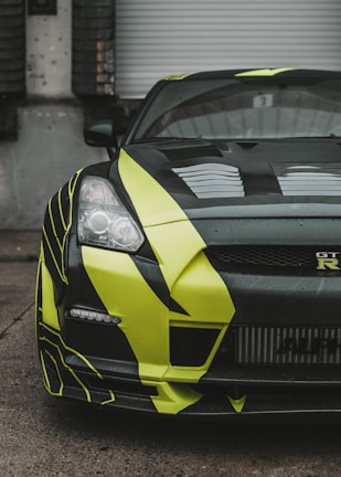 A close-up view of a sports car with a striking black and neon yellow design. The car features sleek, aerodynamic bodywork and is partially parked in front of a gray industrial garage door. The headlights are sharply defined, and the front grille showcases a distinctive logo.