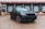A sleek black SUV is parked in front of a loading dock with closed garage doors. The setting is industrial, with a concrete ground and a corrugated metal building. The vehicle has a modern design featuring large wheels and prominent front grilles.