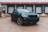 A sleek black SUV is parked in front of a loading dock with closed garage doors. The setting is industrial, with a concrete ground and a corrugated metal building. The vehicle has a modern design featuring large wheels and prominent front grilles.