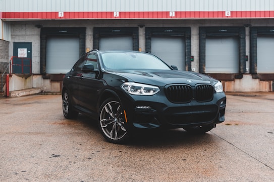 A sleek black SUV is parked in front of a loading dock with closed garage doors. The setting is industrial, with a concrete ground and a corrugated metal building. The vehicle has a modern design featuring large wheels and prominent front grilles.