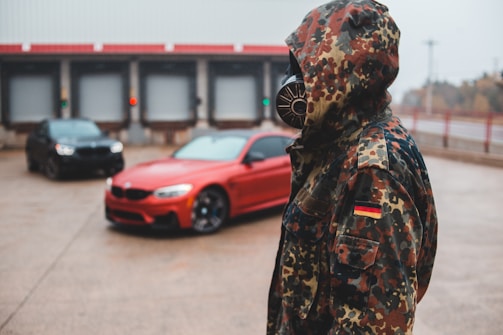 A person in a camouflage jacket with a German flag patch stands facing slightly to the side, wearing a gas mask. In the background, a red car and a darker, possibly black or navy car, are slightly out of focus. The setting appears industrial with warehouse doors visible behind the vehicles.