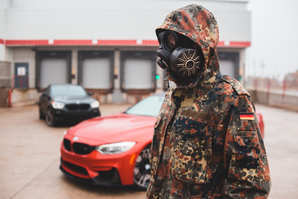 person wearing black and brown floral hoodie and black mask standing beside red car