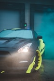 A sporty car with a sleek design and colorful accents is parked in front of large industrial doors, partially enveloped in teal-colored smoke. The car features bright yellow details on the rims and body, with headlights on. The setting appears to be an industrial area, indicated by the metal doors in the background.