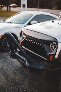 Two cars parked closely next to each other in an outdoor setting. The car in the foreground features a rugged front bumper and distinctive grille design, while the car next to it has a more streamlined appearance. The scene is set on a gravel surface with some greenery and road visible in the background.