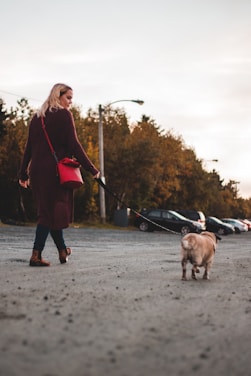 A person with blonde hair walks a small dog on a leash through a parking lot. The individual is wearing a long, dark coat and carrying a red bag. Several cars are parked in the background, and the surroundings are lined with trees under a cloudy sky.