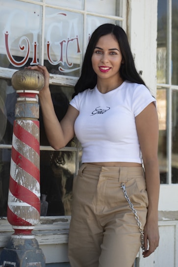 A woman with long dark hair stands beside a vintage barber pole with red, white, and blue stripes. She is wearing a white crop top with a design on it and beige pants with a chain accessory. The background features a window with ornate text painted on it.