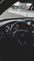 A car interior focused on the steering wheel featuring silver accents and the brand logo. The dashboard displays illuminated gauges with a mix of analog and digital screens. The background shows an urban setting through the windshield.