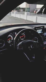 A car interior focused on the steering wheel featuring silver accents and the brand logo. The dashboard displays illuminated gauges with a mix of analog and digital screens. The background shows an urban setting through the windshield.