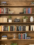 A wooden bookshelf filled with various books arranged neatly on four shelves. Several plants in pots and decorative items, such as a glass dome and candle holders, are interspersed among the books. The bookshelf has a warm, natural wood finish, and the books vary in color and size.