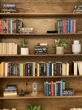 A warm wooden open shelf filled with colorful books and small plants.